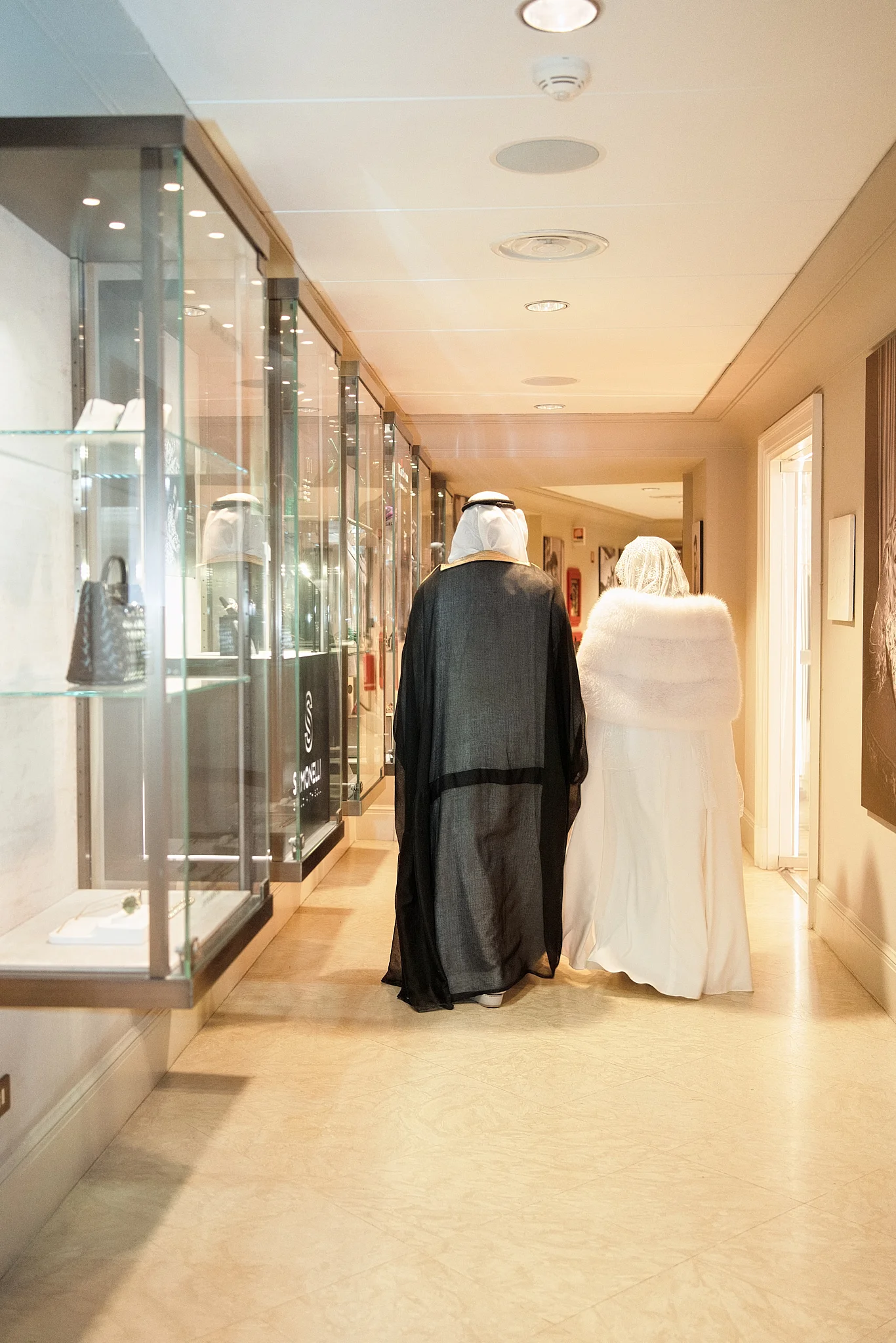 A couple in traditional and white outfits walk down a bright hallway past display cases.
