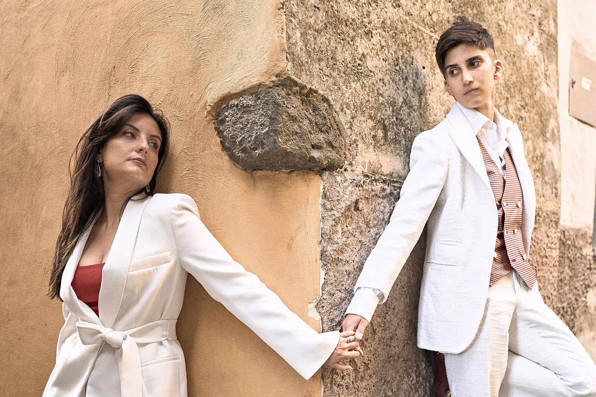 Two women in stylish white suits hold hands around a corner, after getting engaged.
