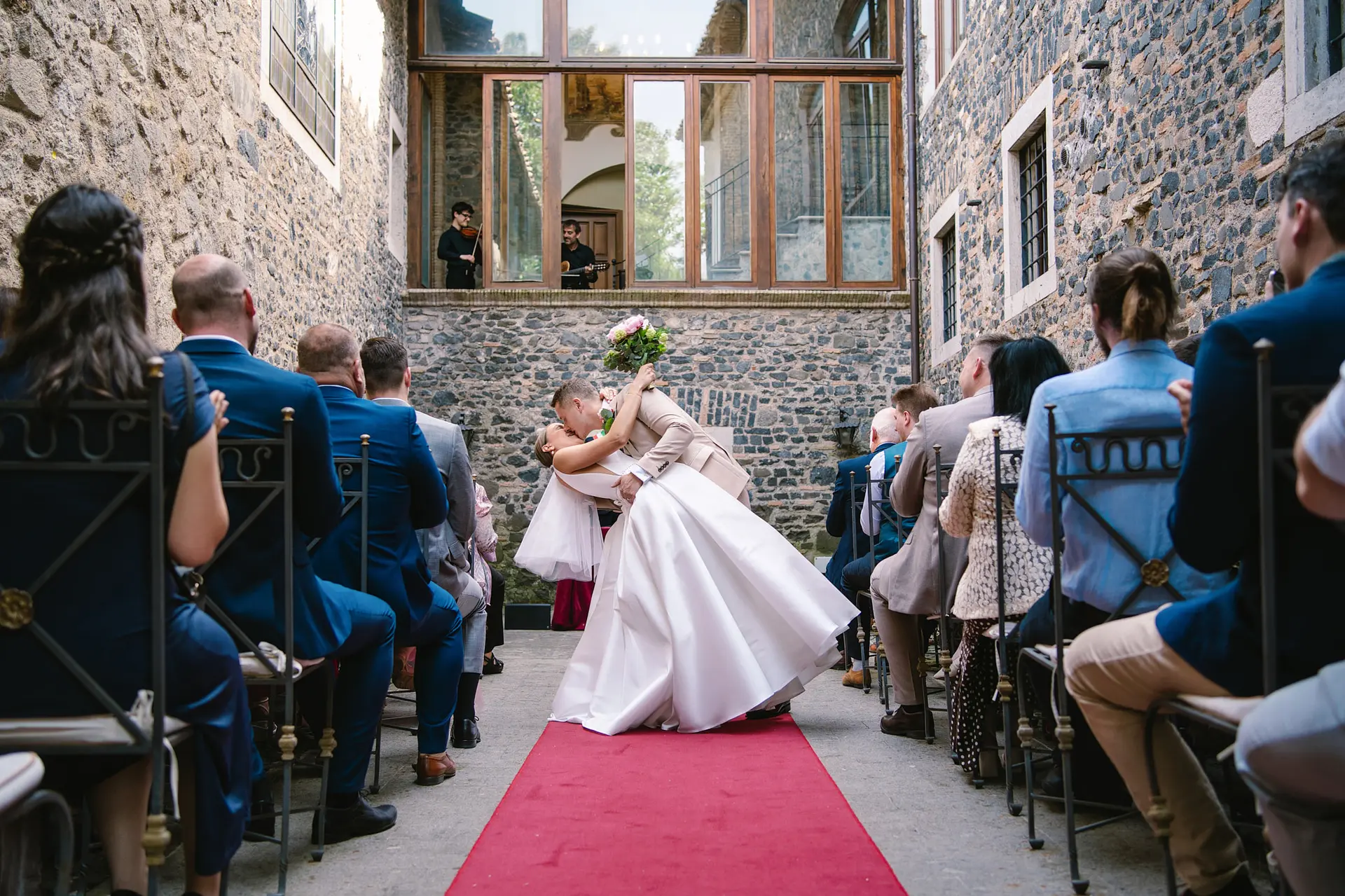 Groom dips bride in a full gown, kissing on a red carpet aisle at their outdoor stone venue wedding.