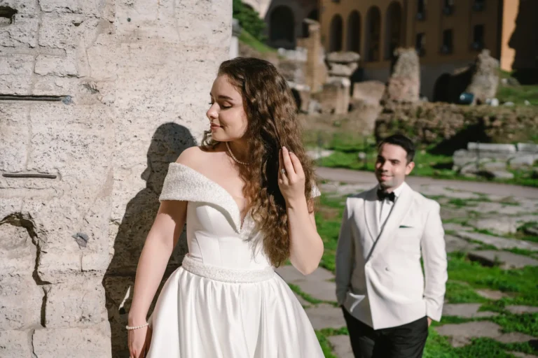 A beautiful bride in a white gown stands by a stone wall in Rome, with the groom smiling behind.