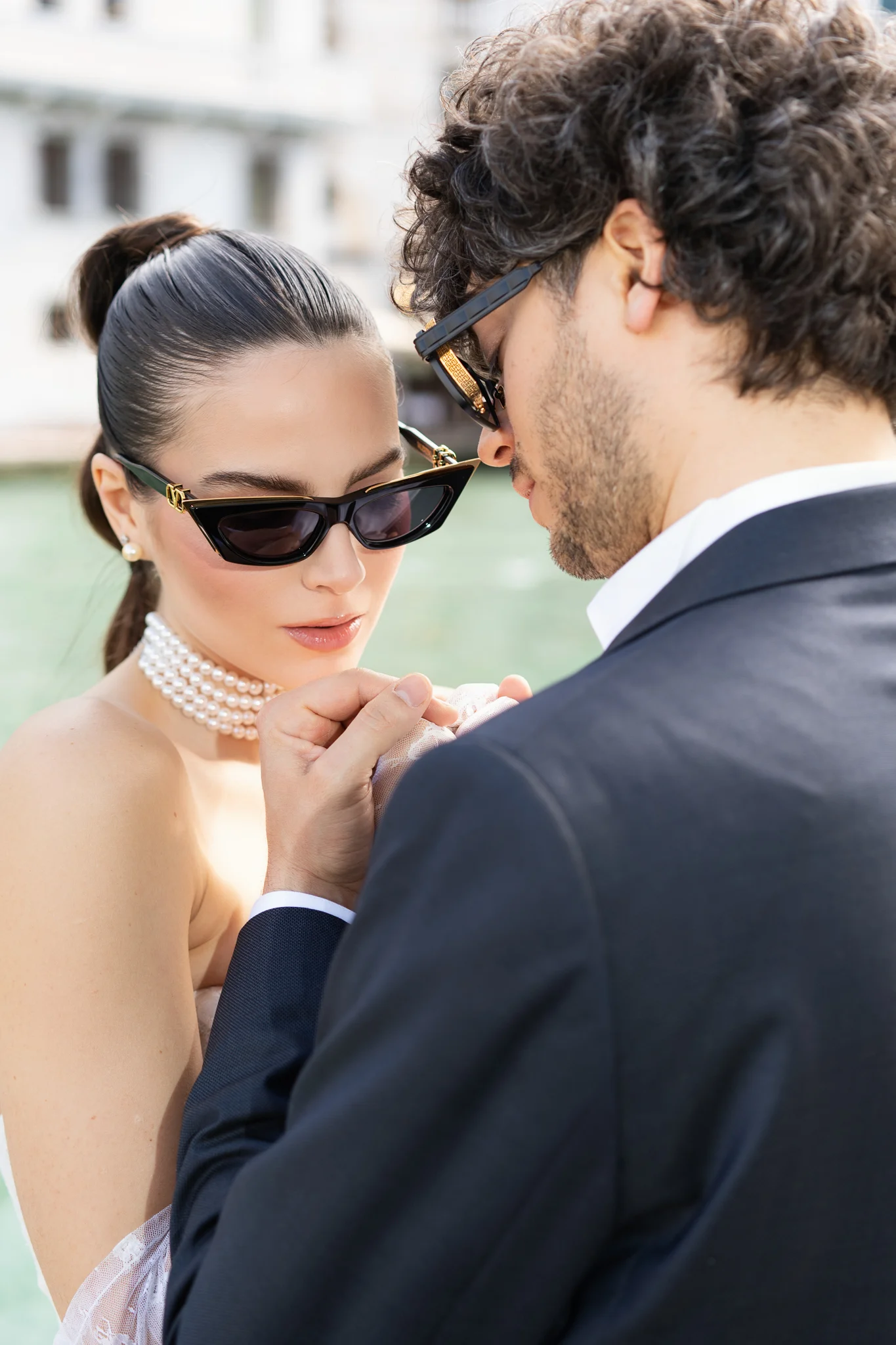 A chic bride in sunglasses and a pearl choker holds hands with her groom in a stylish close-up.