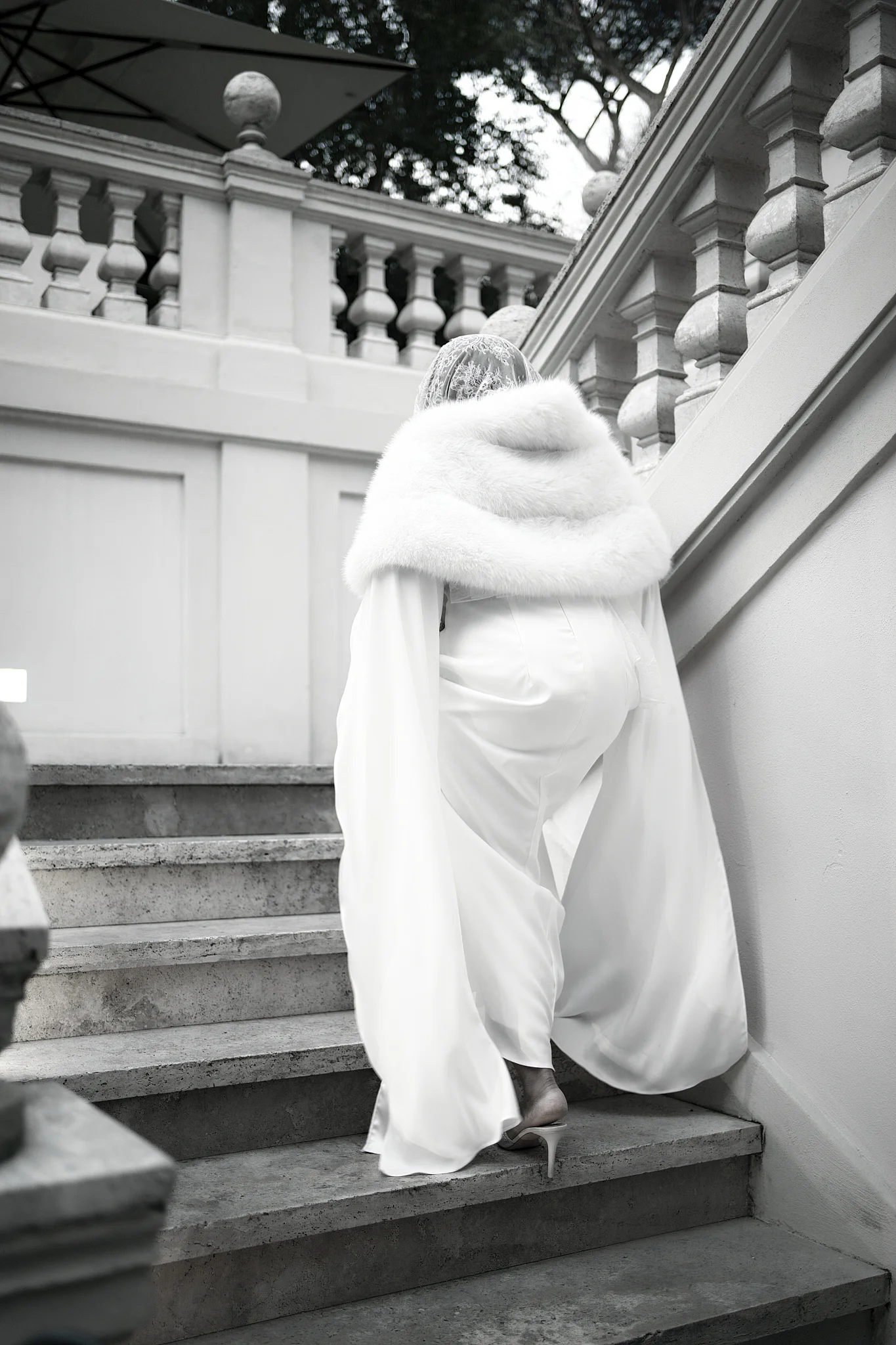 A bride in a chic white jumpsuit with a fur stole ascends a grand stone staircase from behind.