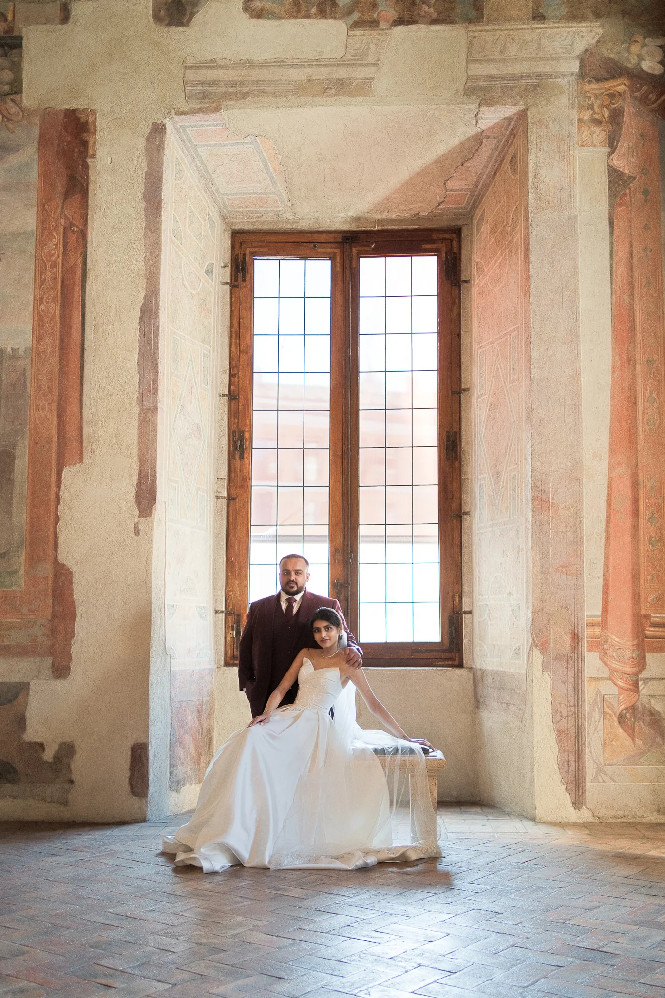 An elegantly dressed couple poses for a portrait by a large window inside a historic Italian villa.