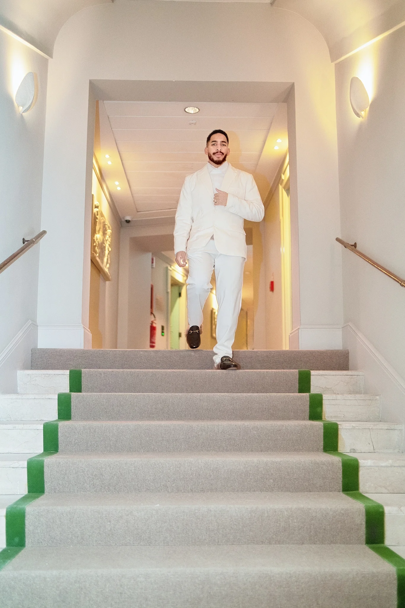 A stylish groom in a white suit and loafers walks down a carpeted staircase with green trim.