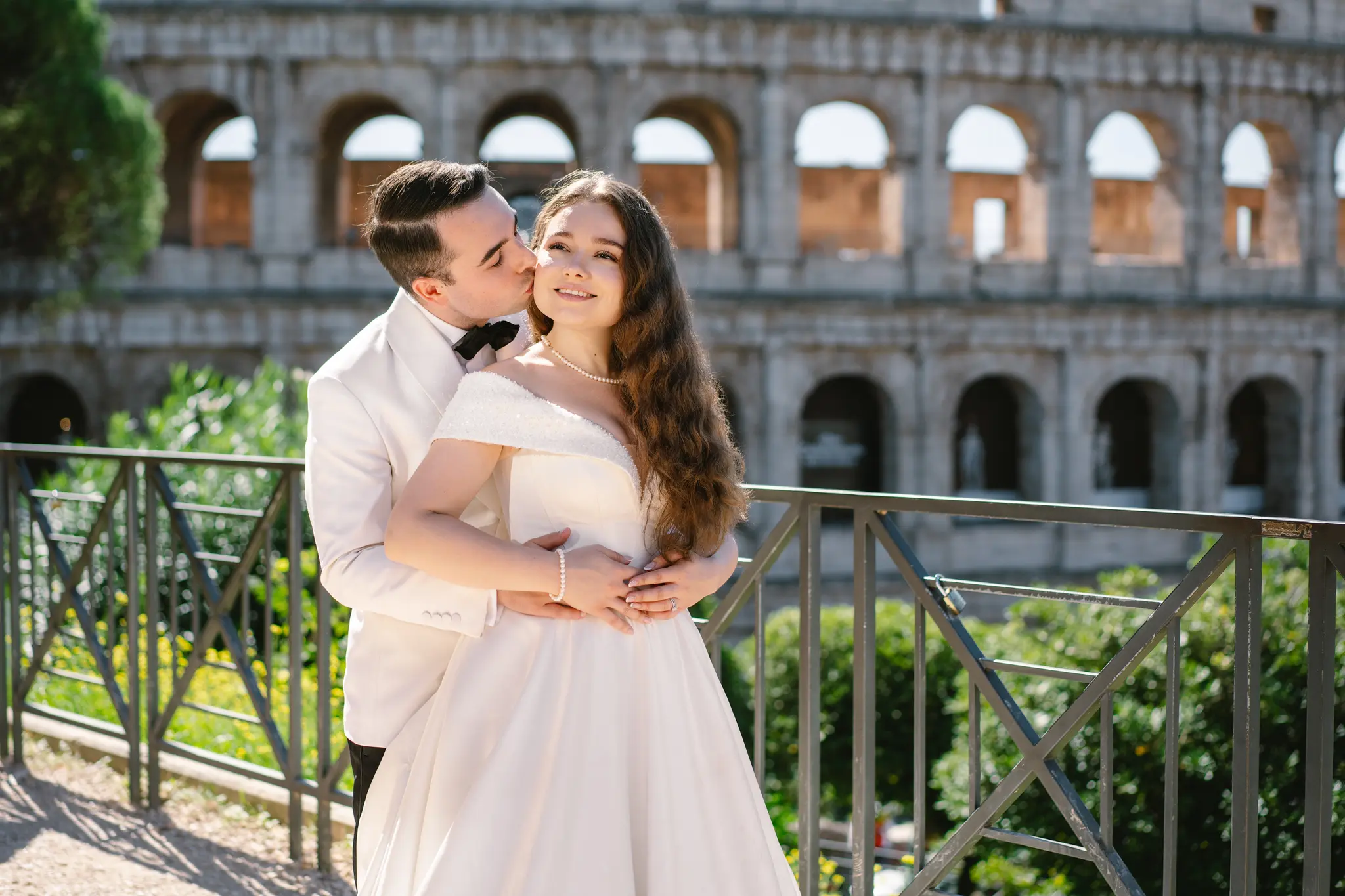 A groom in a white tuxedo tenderly kisses his bride on the cheek, with the iconic Colosseum behind them.