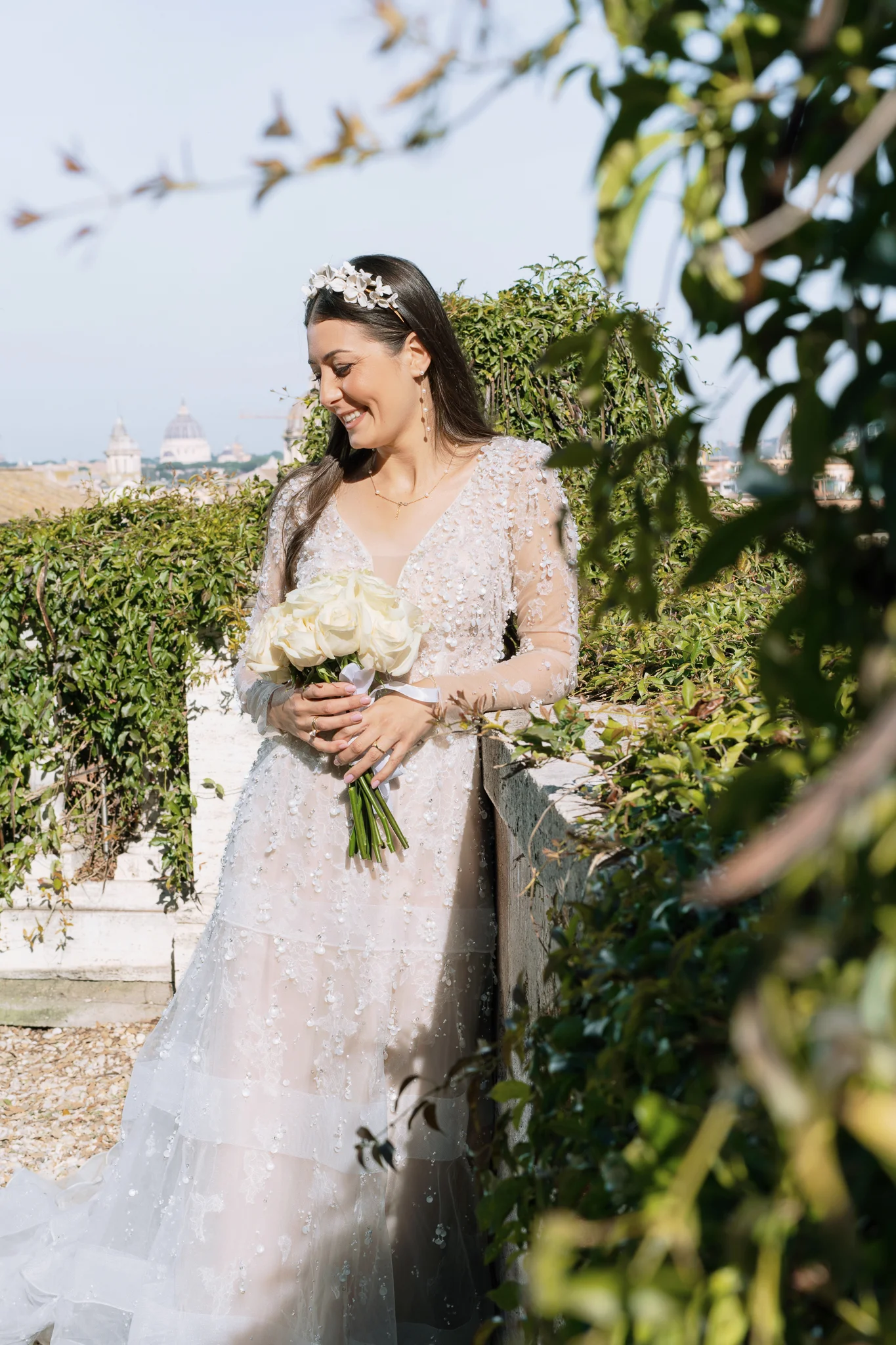 A bride in a stunning long-sleeved dress holds a fresh bouquet of white roses in a garden setting.